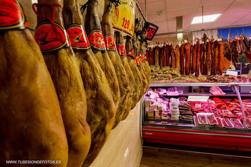 fotografía con jamones colgados y parte de un mostrador con lomos, chorizos, morcillas y salchichón